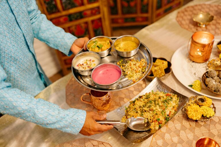 A person serving a traditional Indian thali, featuring a variety of dishes, during a festive indoor dinner setup.