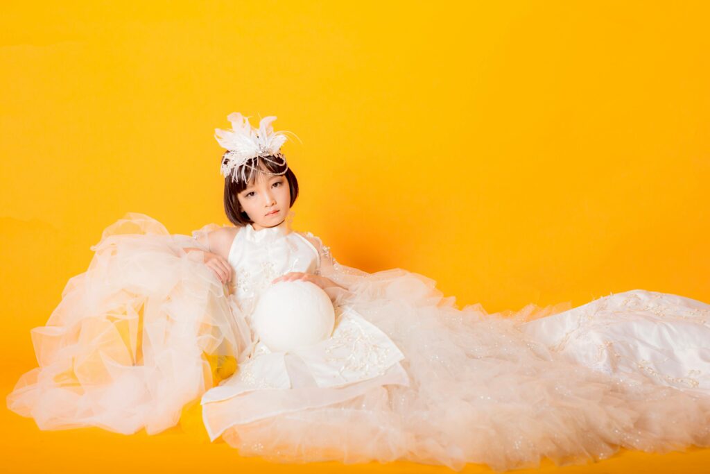 Stylish young girl in an elegant white gown posing against a vibrant yellow background in a creative studio shoot for Children's clothing trends.
